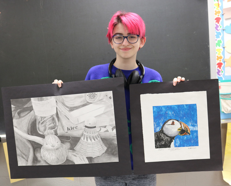 Riley Johnson, senior at Lanesboro High School, poses with her entries for the Juried High School Art Show at Lanesboro Arts. On the left is a still life drawing and on the right is a watercolor print of a puffin. Photo by Barb Jeffers