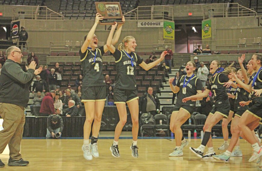 Caledonia seniors Josie Foster (#4) and Sienna Augedahl (#12) hoist the Section 1AA champions trophy while teammates rush in following the Warriors 63-53 win over Dover-Eyota. Caledonia’s girls are off to state. Photo by Paul Trende