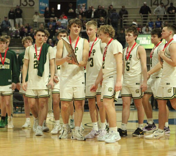 Rushford-Peterson’s lone senior, Caden Johnson holds the Section 1A runner-up trophy as his younger teammates converge. The Trojans trailed most of the section title game versus Goodhue, but made a comeback that fell just short, as the Wildcats prevailed 84-79. Photo by Paul Trende