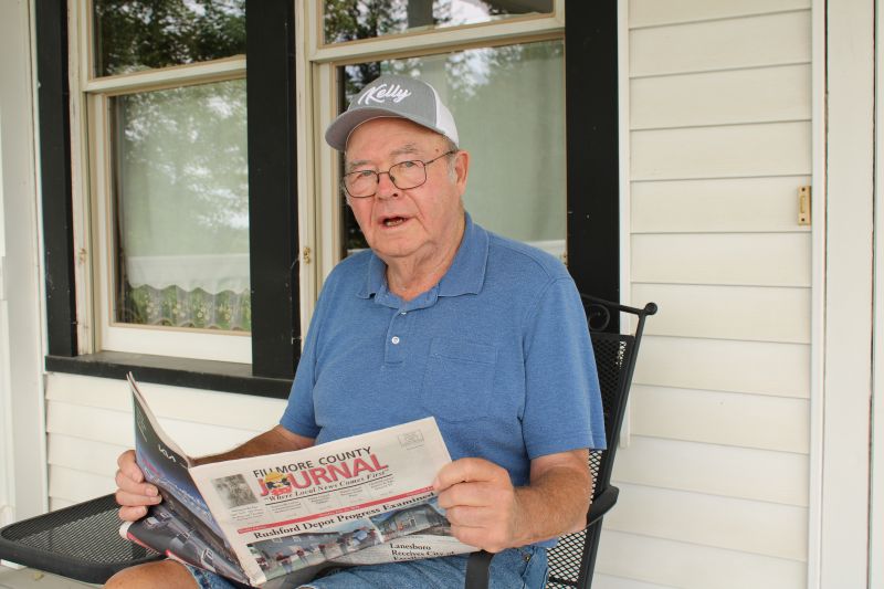 Wearing a hat promoting Kelly Printing & Signs, Richard Kelly looks at a recent issue of the Fillmore County Journal while sitting on the porch of his farmhouse in rural Highland. As he celebrates his 81st birthday, he reflects on a newspaper he and his late wife Esther started half a lifetime ago. One of many successful ventures launched by the Kellys over the past 40 years. Photo by Jason Sethre